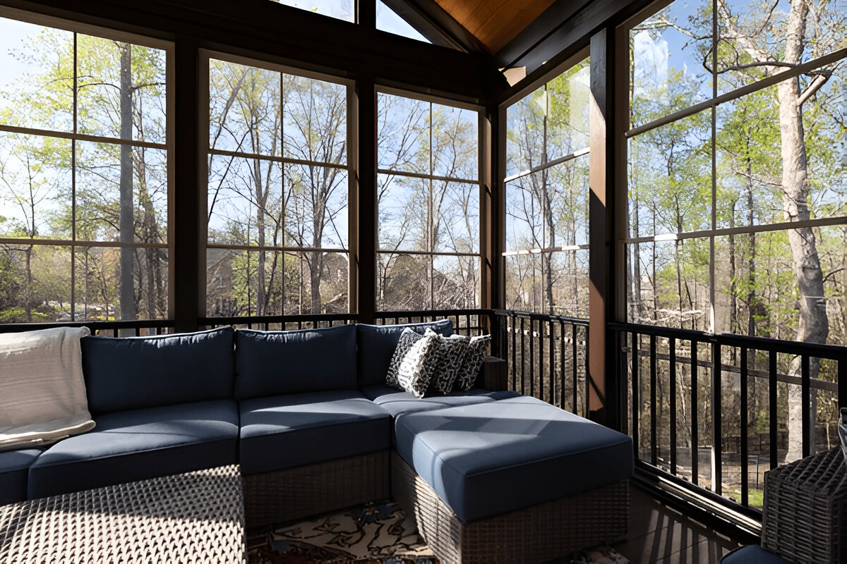 Cozy sunroom with large screened windows, dark wood framing, and a blue cushioned sectional sofa overlooking a wooded view.