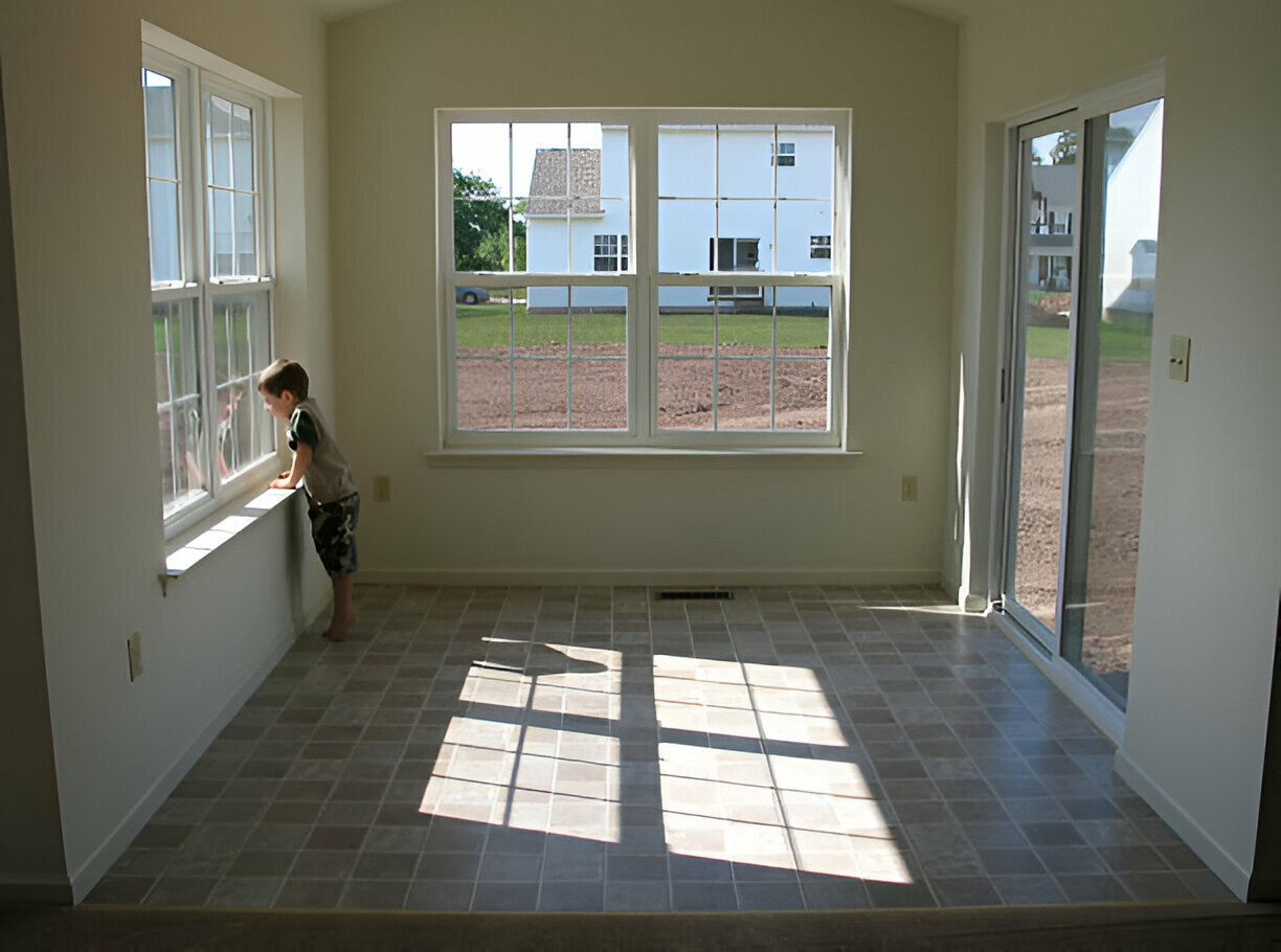 
Child standing by the window in a bright, empty sunroom with tile flooring and large windows