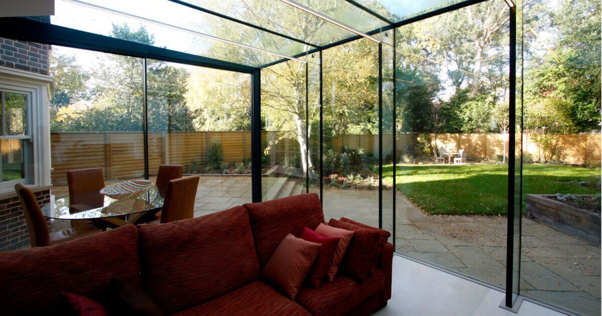 Glass sunroom extension with floor-to-ceiling windows and a clear roof, featuring a red couch and backyard garden view.