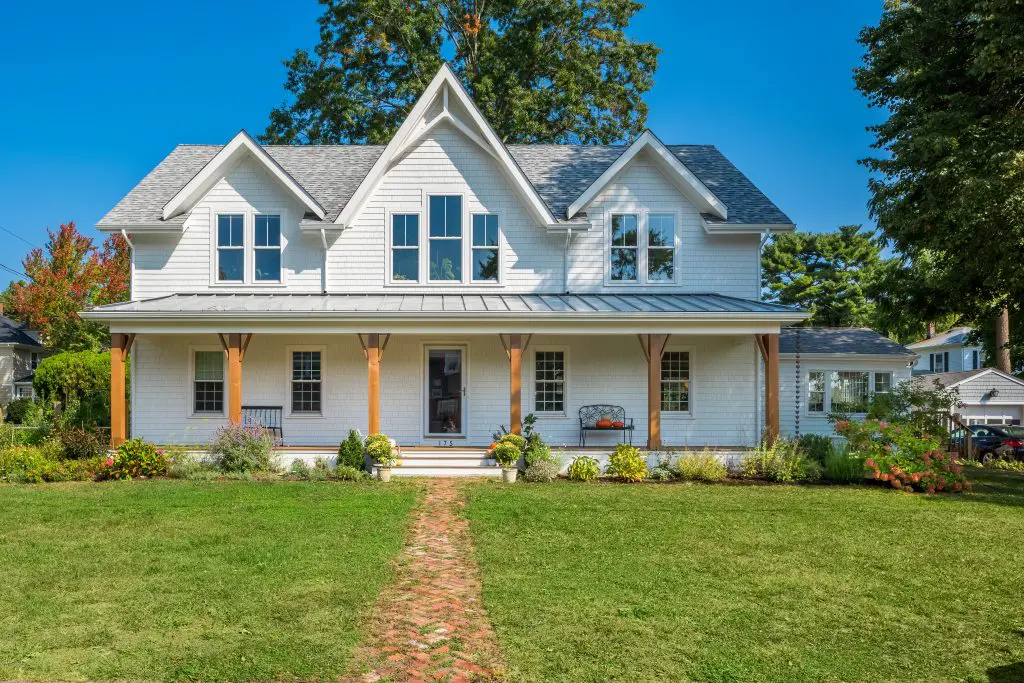  The image shows a beautiful two-story white farmhouse-style home with a wraparound porch, multiple gabled roofs, and large windows. The home is surrounded by a lush, well-maintained lawn with a brick pathway leading to the front door. The exterior features white siding, dark-colored trim, and a metal roof, creating a classic and timeless aesthetic. The surrounding trees and landscaping add to the picturesque and inviting atmosphere