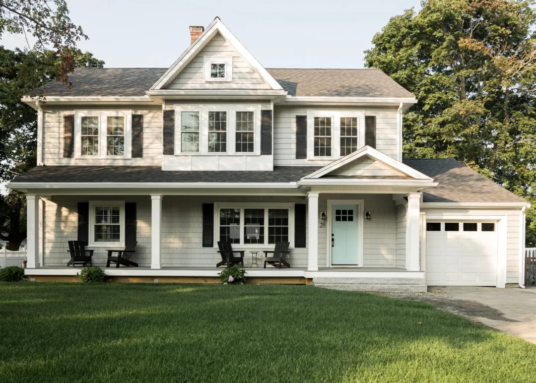  The image shows a beautiful two-story farmhouse-style home with a wraparound porch, multiple gabled roofs, and large windows. The home is surrounded by a well-maintained lawn and mature trees, creating a picturesque and inviting setting. The exterior features white siding, dark-colored trim, and a gray asphalt shingle roof, giving the home a classic and timeless aesthetic.