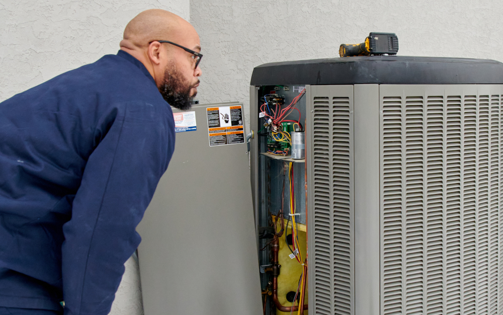  The image shows a male HVAC (Heating, Ventilation, and Air Conditioning) technician in a blue uniform closely examining the internal components of an outdoor air conditioning unit. The technician appears to be troubleshooting or performing maintenance on the unit, likely checking for any issues or ensuring proper functioning