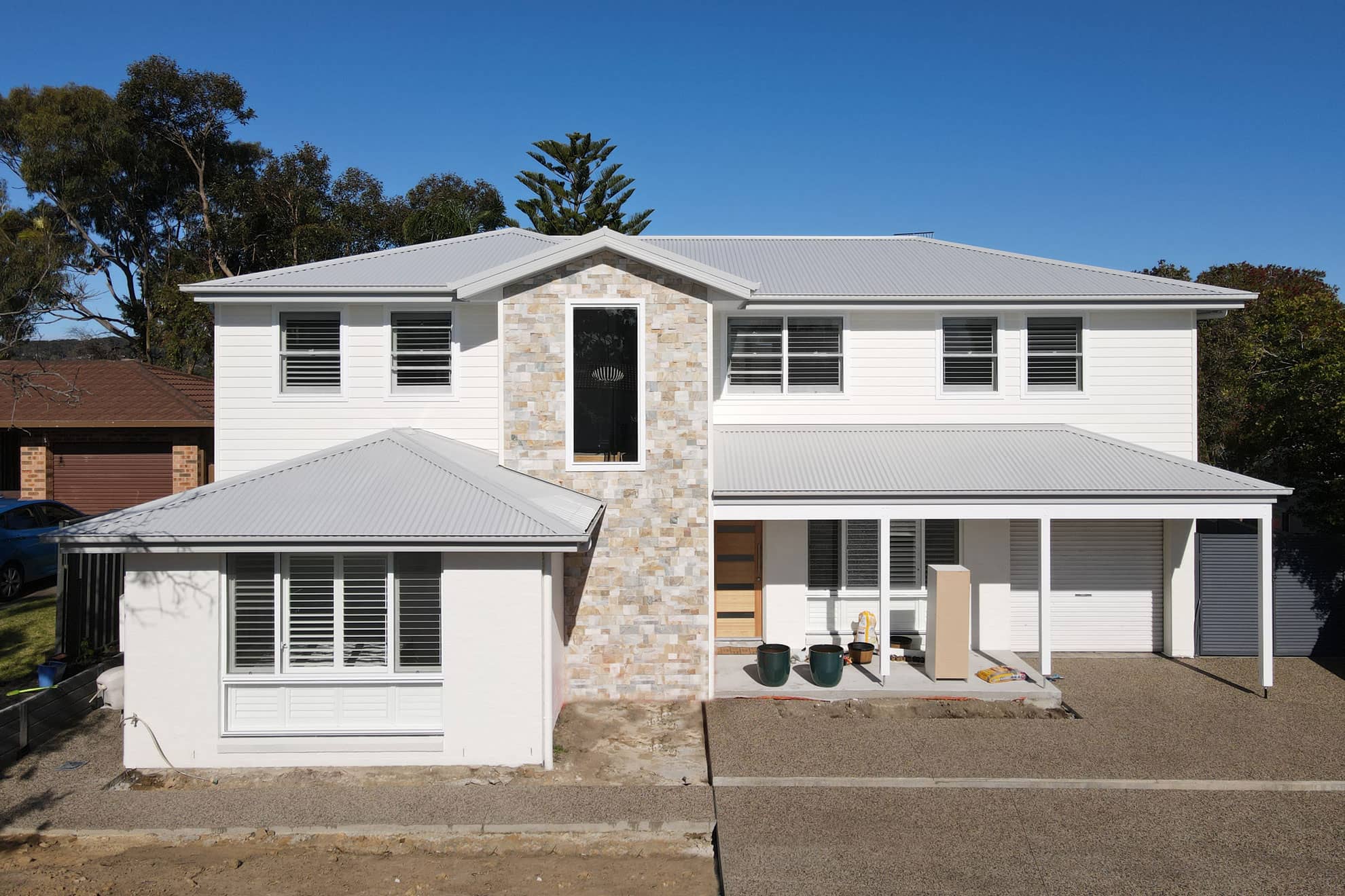 The image shows a two-story, white-painted house with a mix of stone and siding exterior. The house has a wide, covered porch, a gabled roof, and large windows. There are palm trees and other lush vegetation surrounding the property, creating a coastal, tropical atmosphere.
