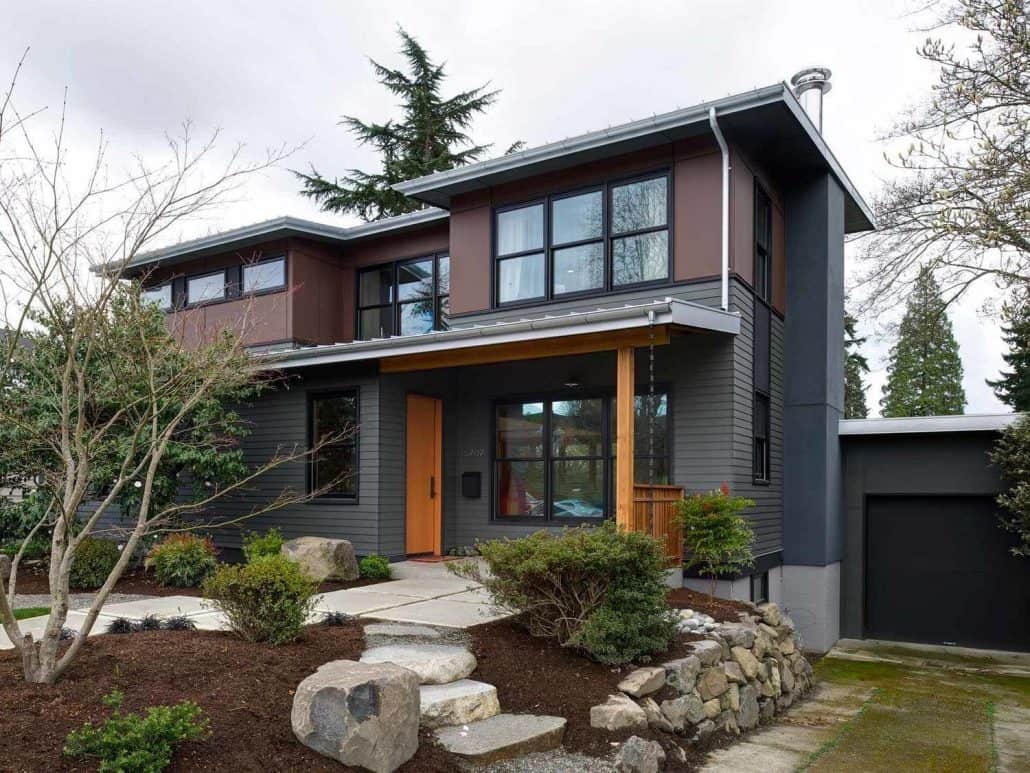 The image shows a two-story home with a modern, farmhouse-inspired design. The exterior features a mix of dark siding, large windows, and a prominent roofline with a gable. The home is surrounded by a well-landscaped yard with stone pathways, shrubs, and a mature tree. The overall aesthetic is a blend of contemporary and traditional elements, creating a visually appealing and harmonious design.