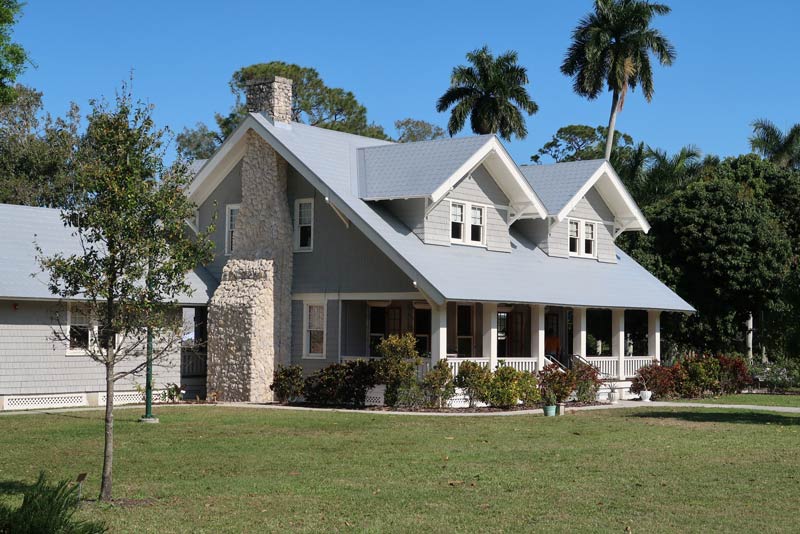 The image shows a large, two-story house with a distinctive tropical-style architecture. The house has a wide, wraparound porch, a stone chimney, and a mix of gabled and hipped roofs. The exterior is painted in a light gray color, and the roof is covered in dark shingles. There are palm trees and other lush vegetation surrounding the house, creating a picturesque, tropical setting.