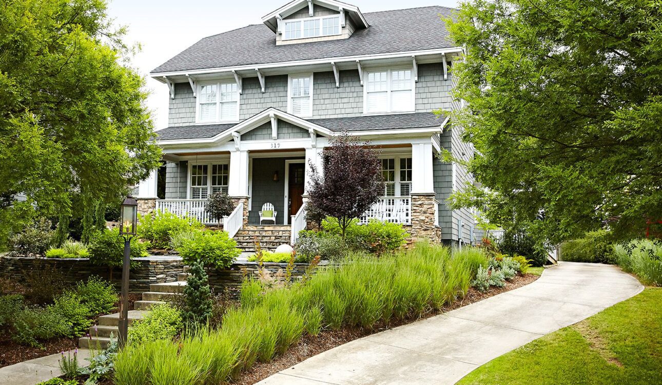 The image shows a two-story Craftsman-style house with a gabled roof, wide front porch, and stone accents. The house is surrounded by a well-manicured landscape, including a paved walkway, lush greenery, and flowering plants