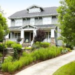 The image shows a two-story Craftsman-style house with a gabled roof, wide front porch, and stone accents. The house is surrounded by a well-manicured landscape, including a paved walkway, lush greenery, and flowering plants
