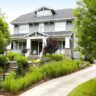 The image shows a two-story Craftsman-style house with a gabled roof, wide front porch, and stone accents. The house is surrounded by a well-manicured landscape, including a paved walkway, lush greenery, and flowering plants