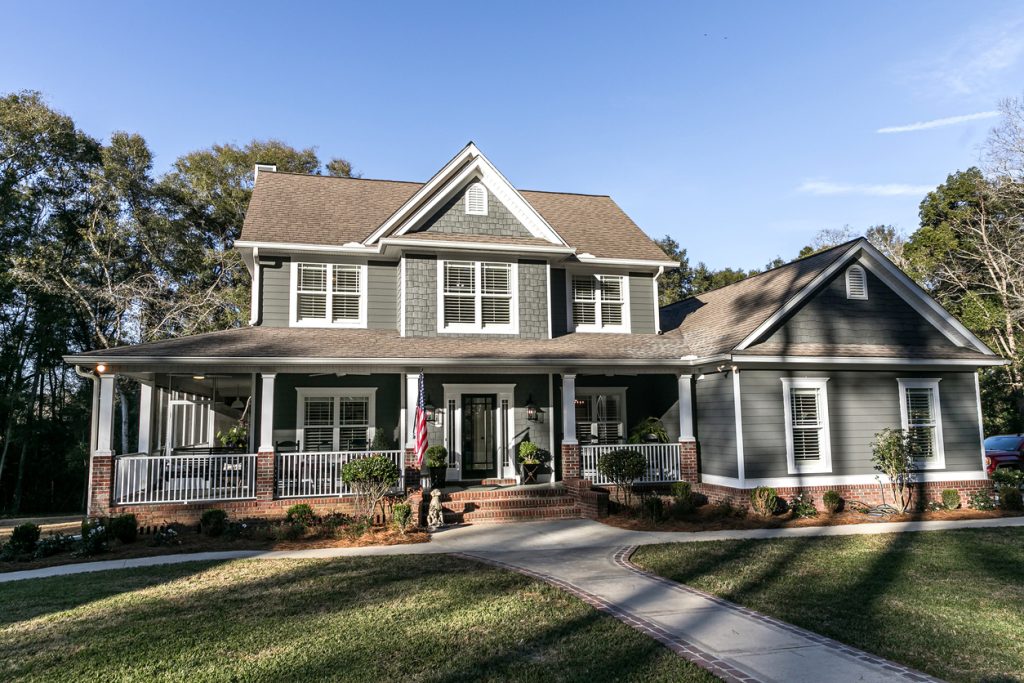 The image shows a large, two-story house with a wraparound porch, gabled roof, and dormers. The exterior is a mix of siding and brick, with a prominent chimney. The house is surrounded by lush, mature trees and landscaping, giving it a traditional, country-style appearance