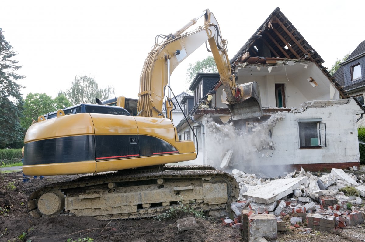  The image shows a large excavator or demolition machine in the process of tearing down a building. The machine is equipped with a powerful arm and bucket, which is being used to break apart the structure. Debris and rubble from the demolished building can be seen scattered around the site, indicating that the demolition process is well underway.