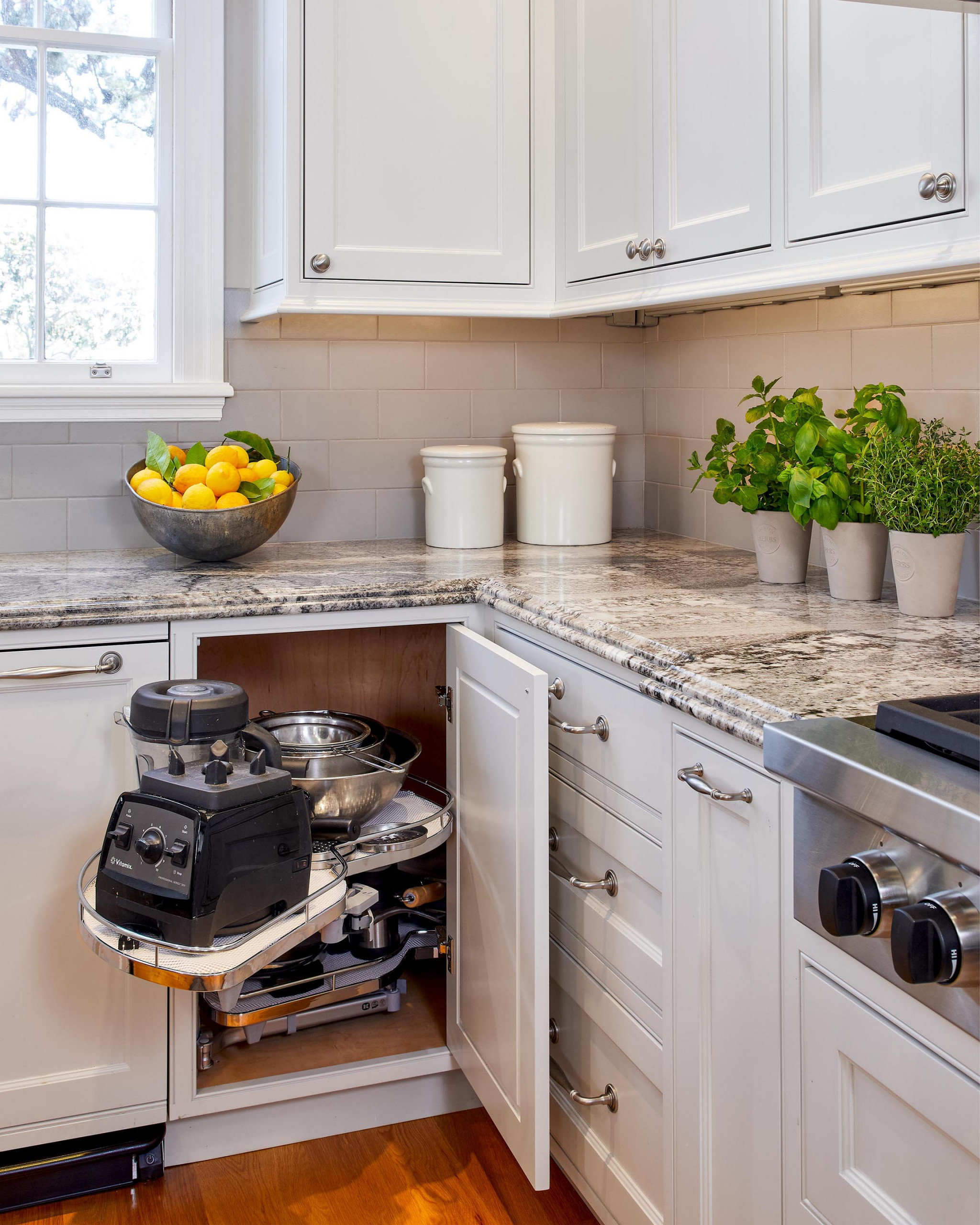 White corner kitchen cabinet with pull-out shelf holding blender and mixing bowls, granite countertops, and herb pots