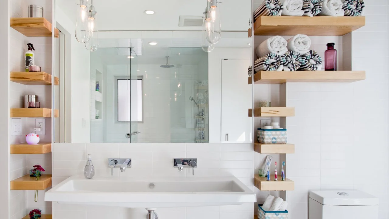 Contemporary white bathroom featuring a double sink, glass shower, and natural wood open shelving with rolled towels and toiletries