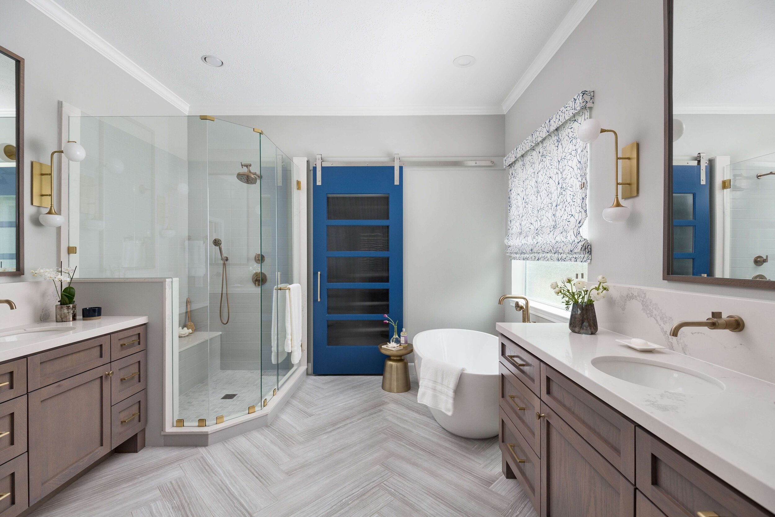 Modern master bathroom featuring a freestanding tub, dual vanities, glass-enclosed shower, and a bold blue barn door