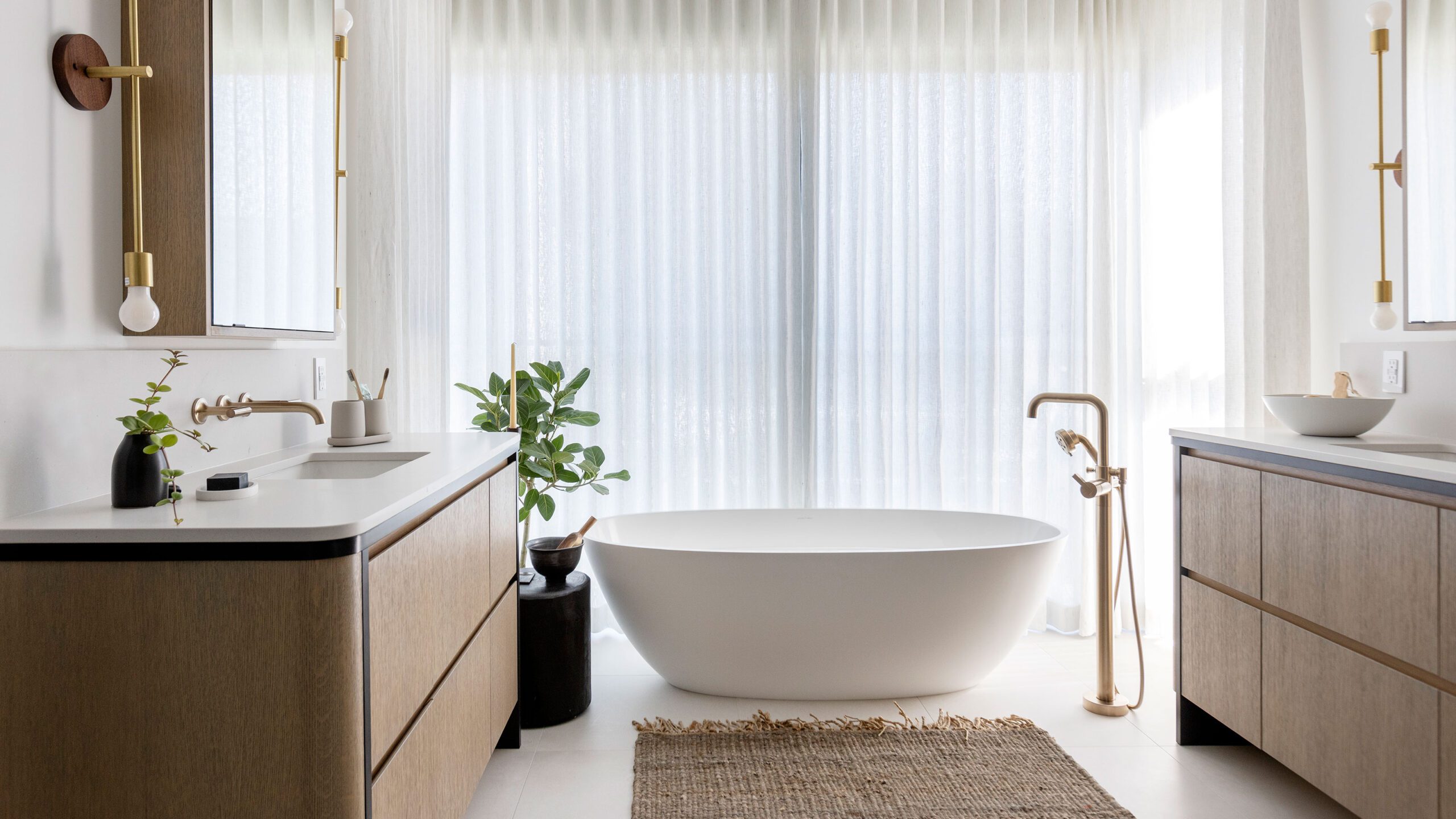 Bright minimalist bathroom featuring a white freestanding tub, wooden vanities, soft lighting, and sheer curtains.