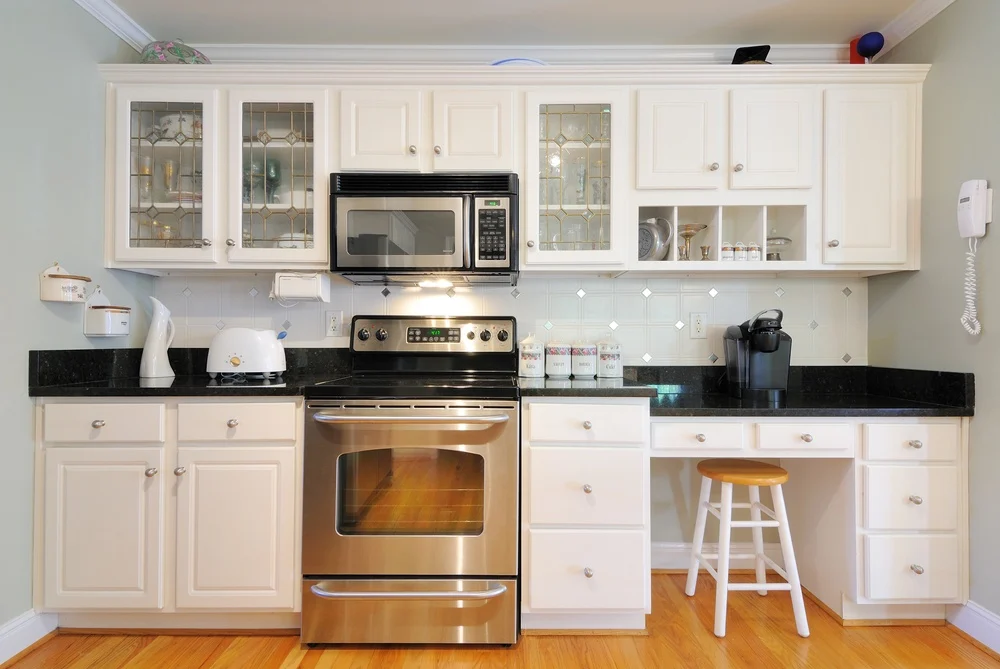 Traditional kitchen featuring white cabinets with glass inserts, stainless steel appliances, black granite countertops, and built-in desk area