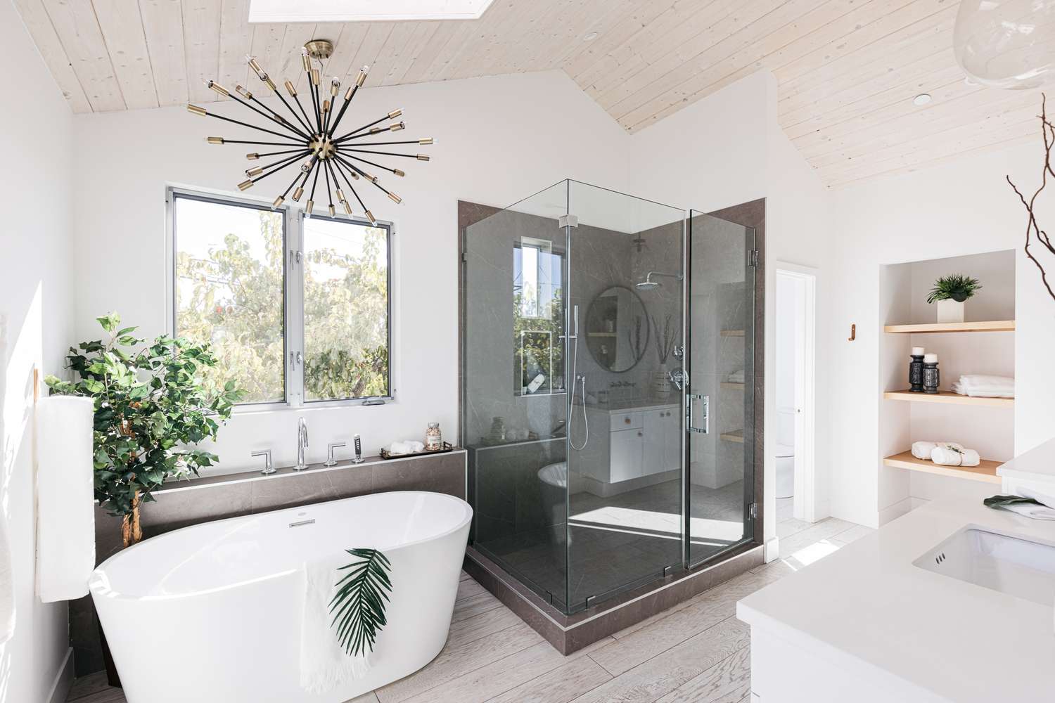 Luxury white bathroom with a freestanding tub, glass-enclosed shower, vaulted ceiling, and modern chandelier.