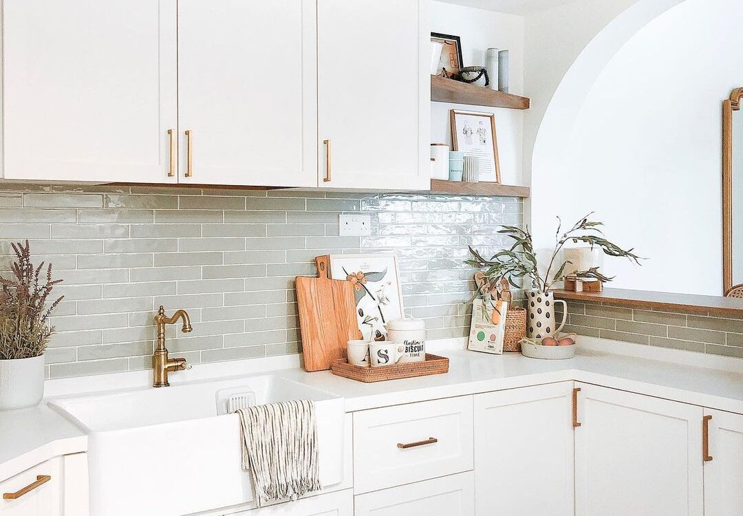 Farmhouse-style kitchen with white shaker cabinets, brass hardware, a farmhouse sink, light green subway tile backsplash, and wooden accents.