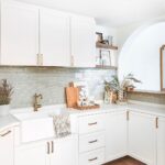 Farmhouse-style kitchen with white shaker cabinets, brass hardware, a farmhouse sink, light green subway tile backsplash, and wooden accents.