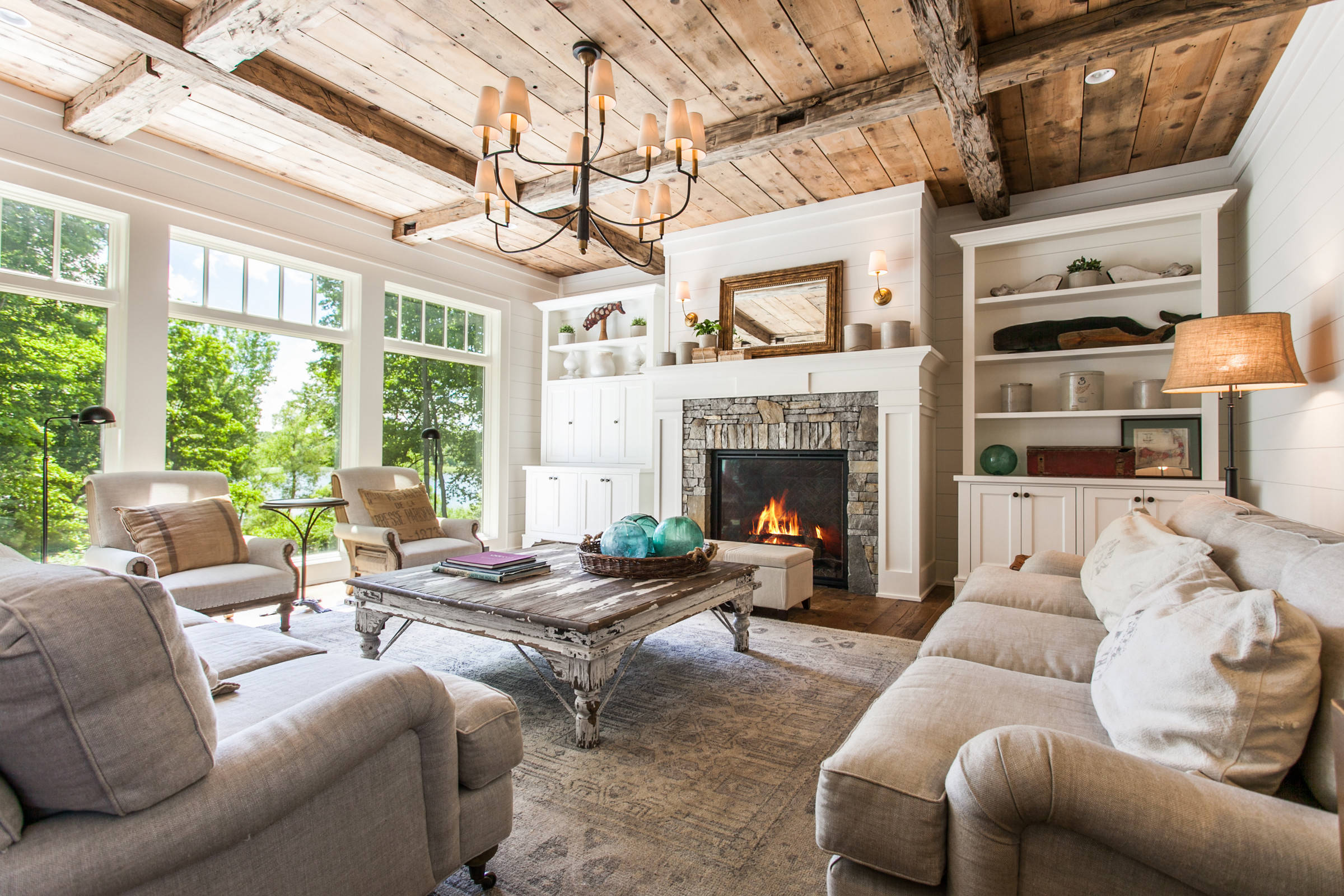 Cozy living room with large windows, rustic wood beam ceiling, stone fireplace, beige upholstered sofas, and a distressed wooden coffee table.