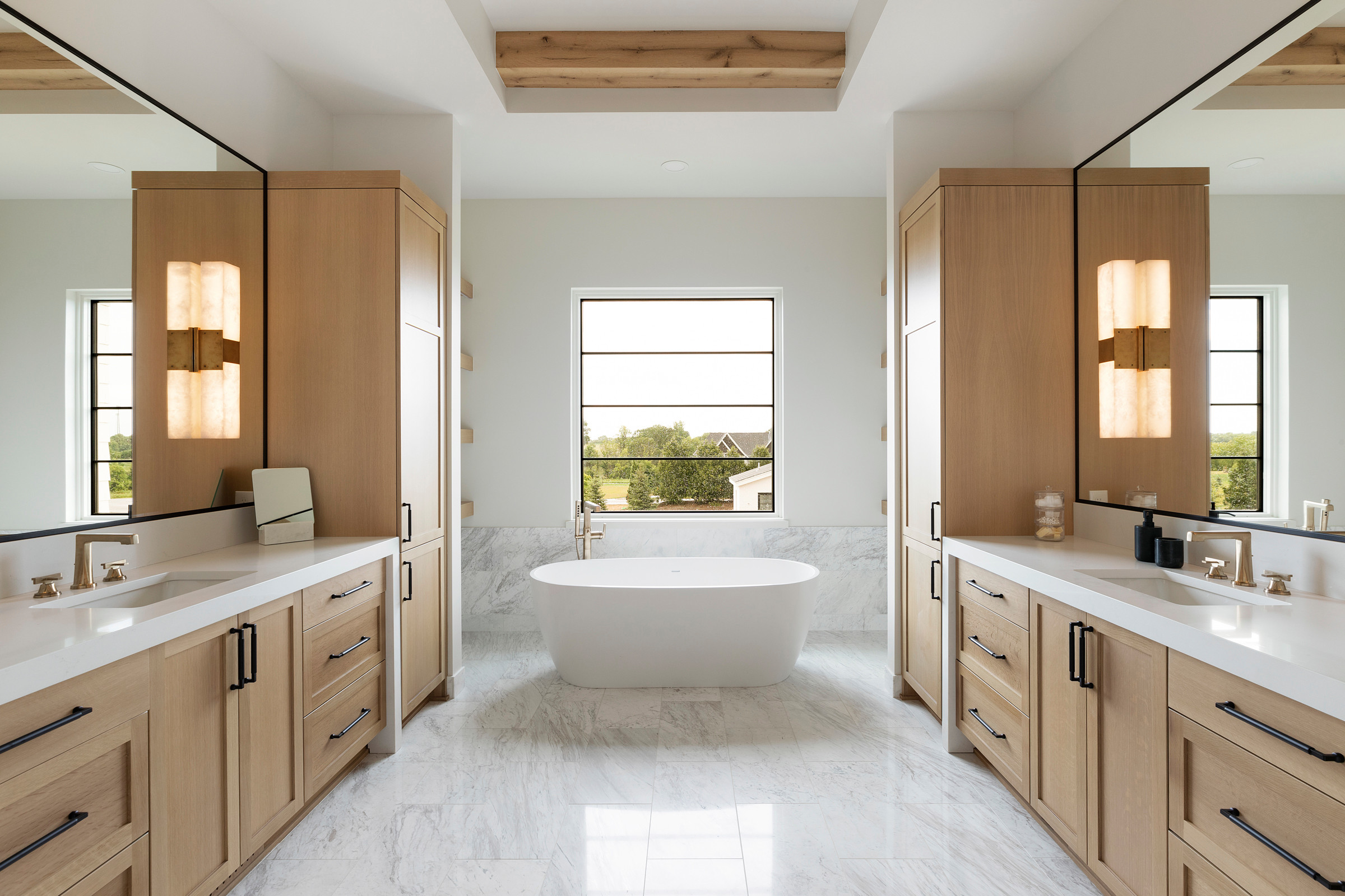 Modern bathroom with freestanding tub, light wood cabinets, dual sinks, and marble flooring
