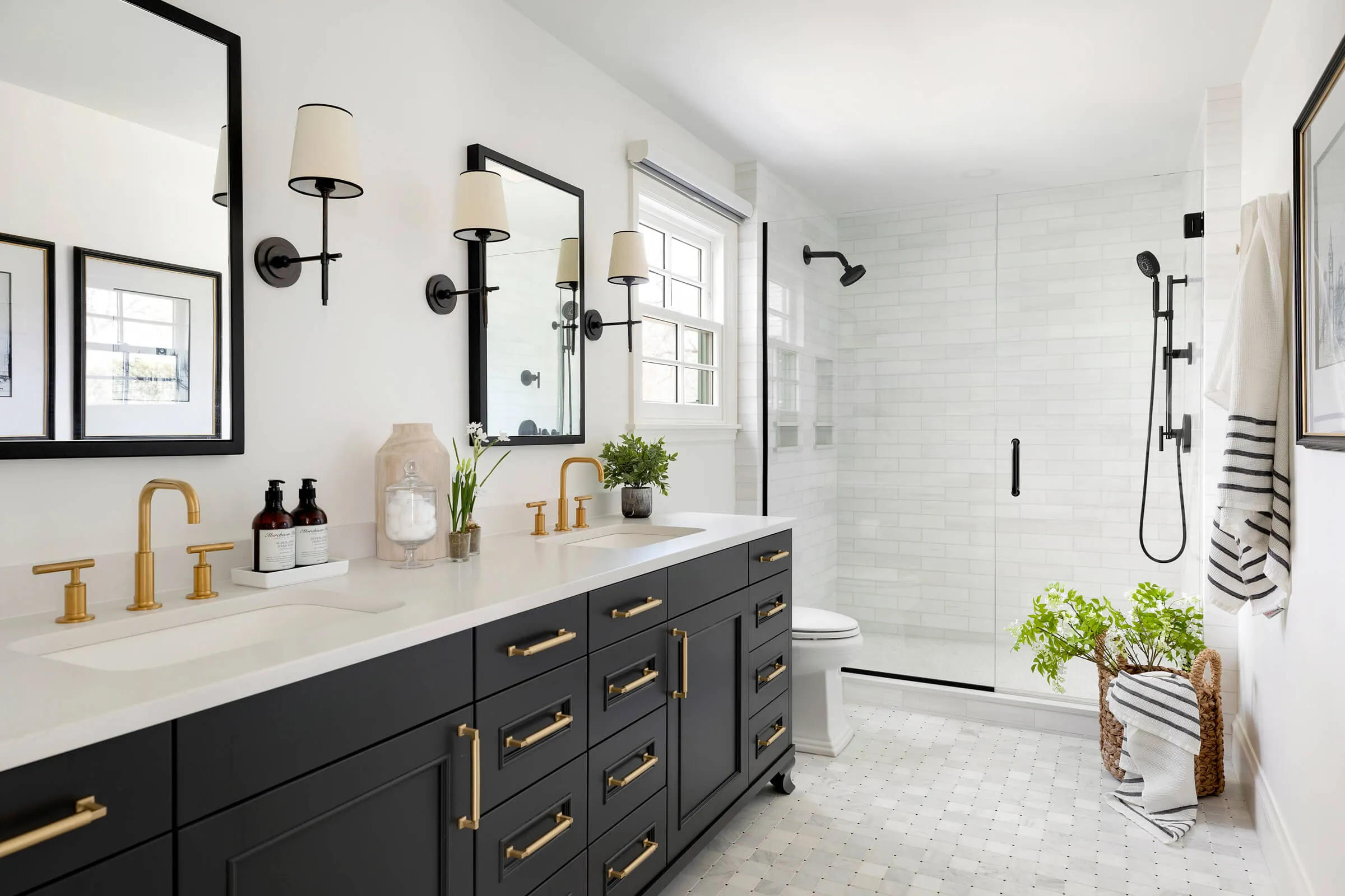 Bathroom featuring a black vanity with gold hardware and faucets, dual sinks, framed mirrors, a glass walk-in shower, and white subway tile walls.