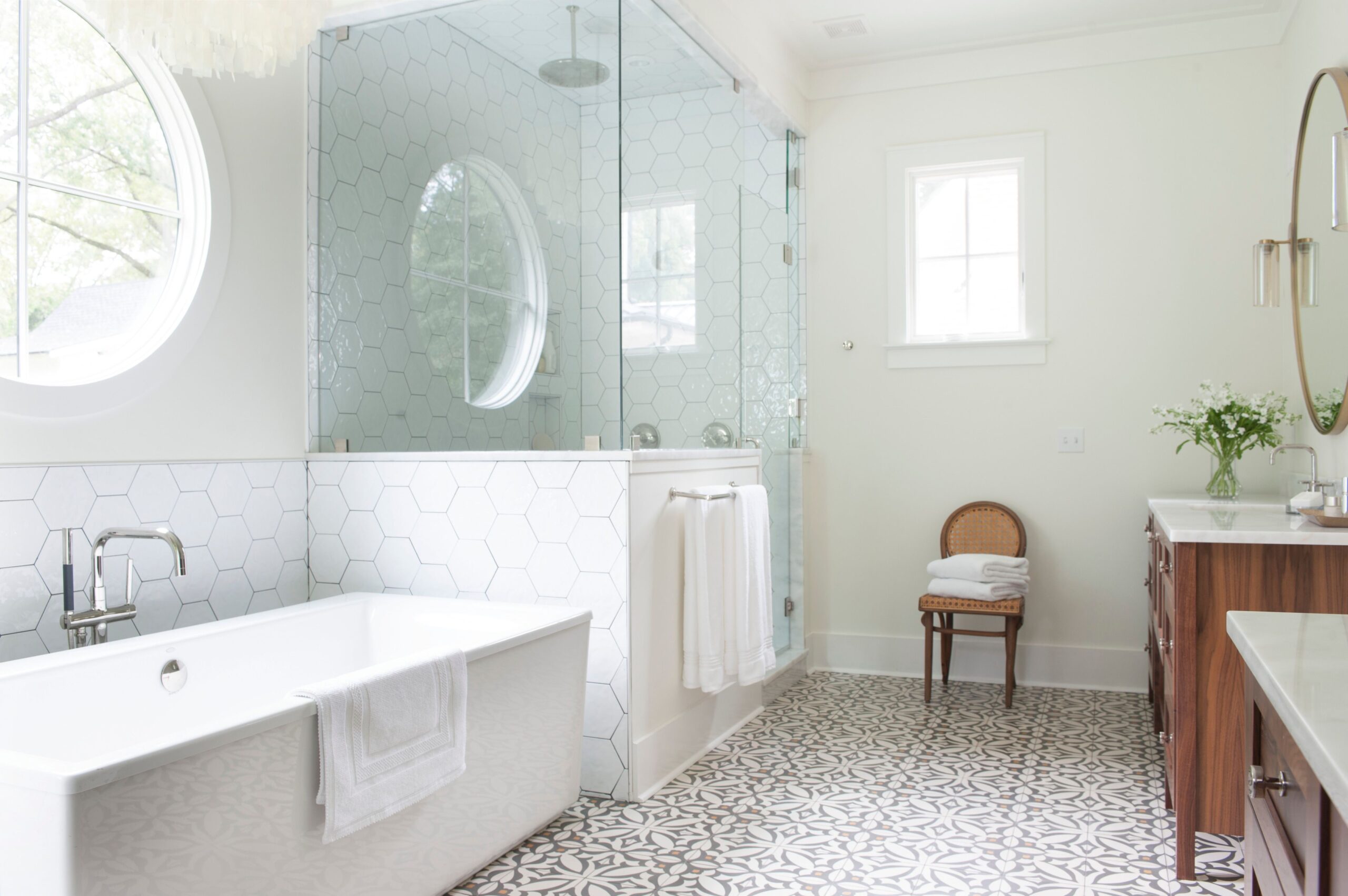 Bright bathroom with freestanding tub, glass shower enclosure, hexagon tile wall, patterned floor, and wooden vanity.