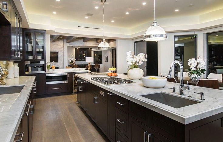 Spacious modern kitchen featuring dark wood cabinets, sleek marble countertops, stainless steel appliances, and two large islands with integrated sink and cooktop.