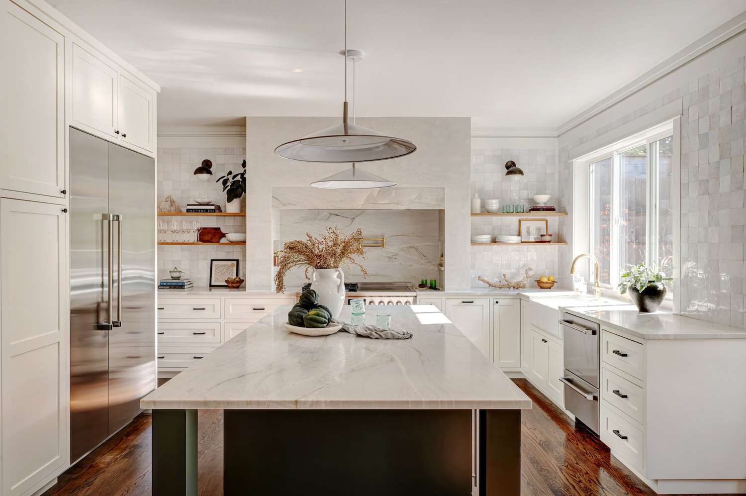 Bright kitchen featuring white cabinets, marble countertops, stainless steel appliances, open wooden shelves, and large pendant lights.