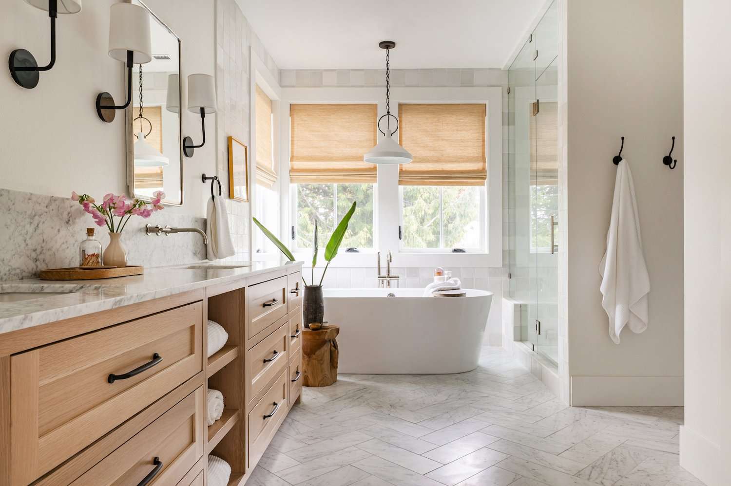 Bright bathroom with freestanding bathtub, light wood vanity, marble finishes, and large windows.