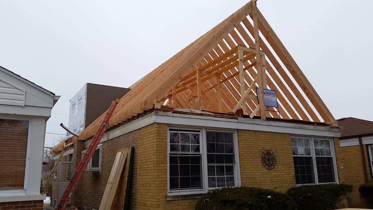  A single-story brick house with exposed wooden trusses showing the framing of a new second-story addition.