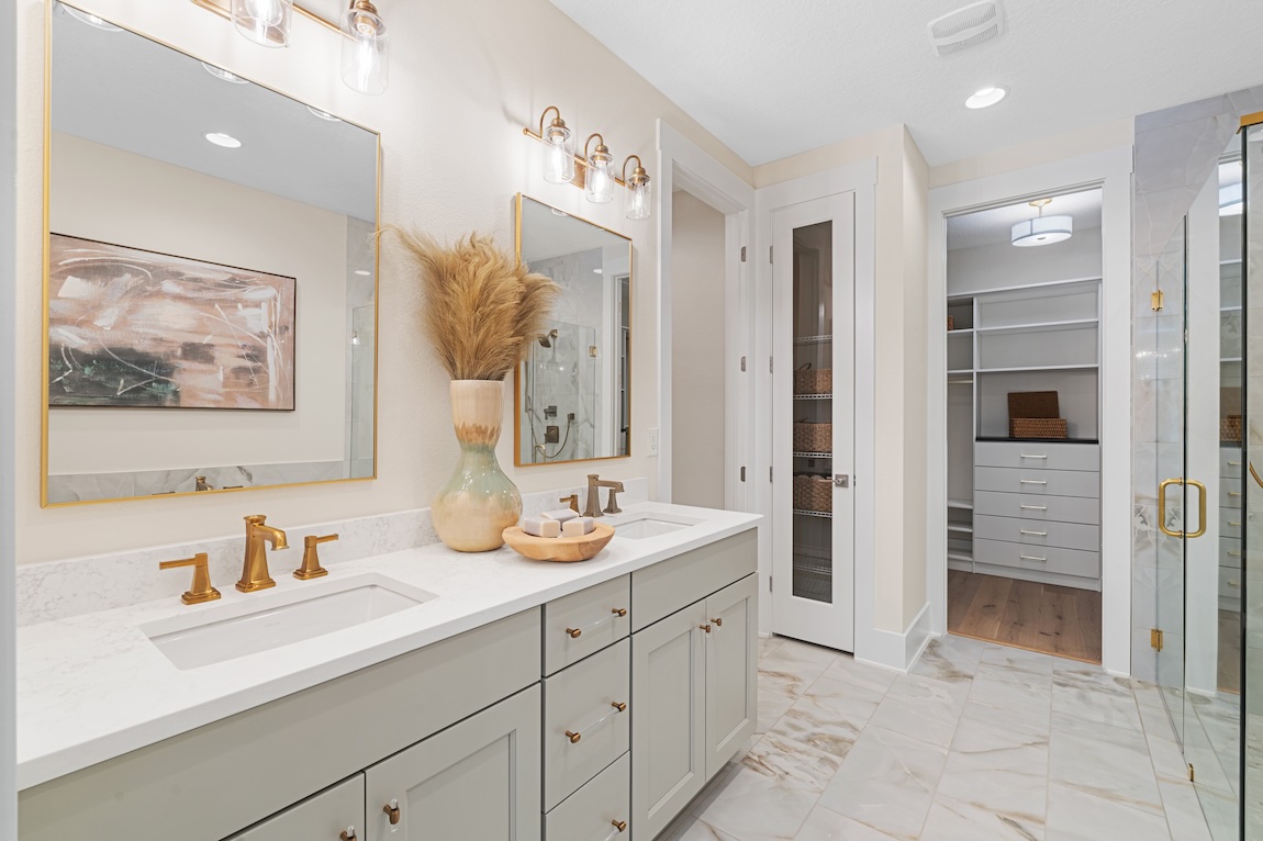 Elegant bathroom featuring a double vanity with gold fixtures, marble flooring, glass shower enclosure, and access to a spacious walk-in closet.