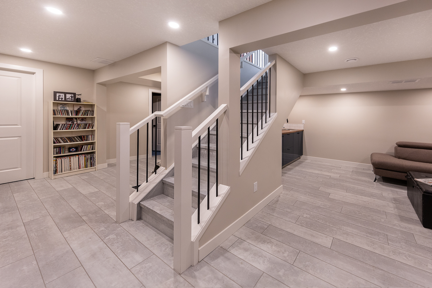 Renovated basement featuring light gray tile flooring, white stair railing with black spindles, bookshelf, and a cozy lounge corner.