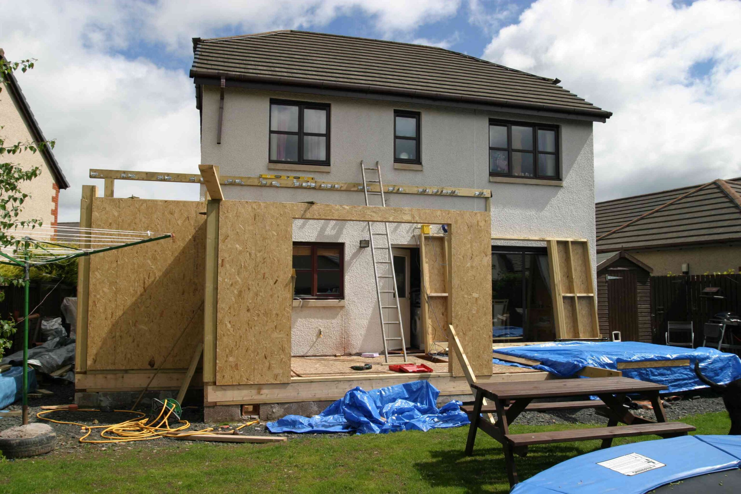 A house undergoing a rear extension with wooden framing and construction materials spread around the backyard.
