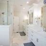 Elegant white bathroom with glass shower, double vanities, and gold accents.
