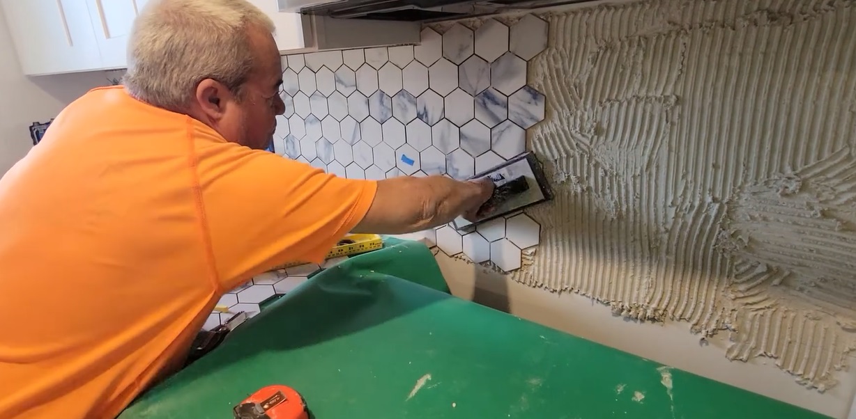 Contractor installing white and gray hexagon tile backsplash with adhesive in a kitchen renovation project.