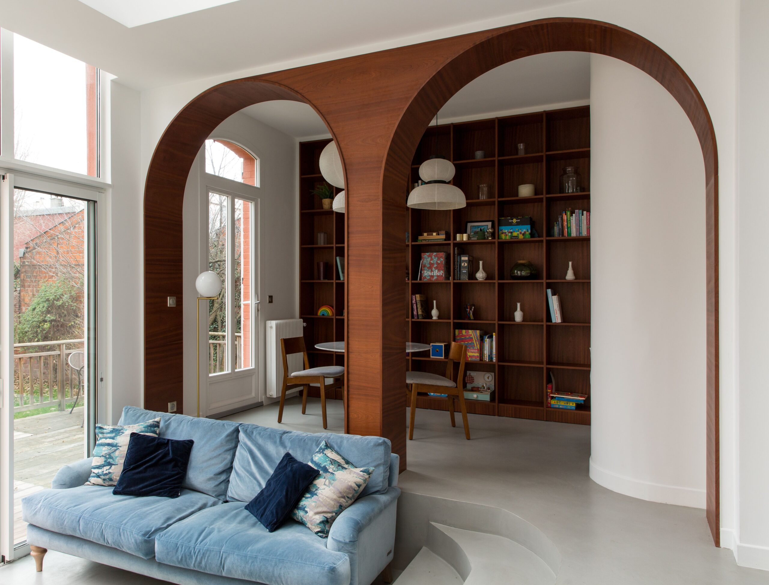 Living area featuring elegant wooden archways, a cozy blue sofa, built-in wood bookshelves, and a small reading nook with natural light.