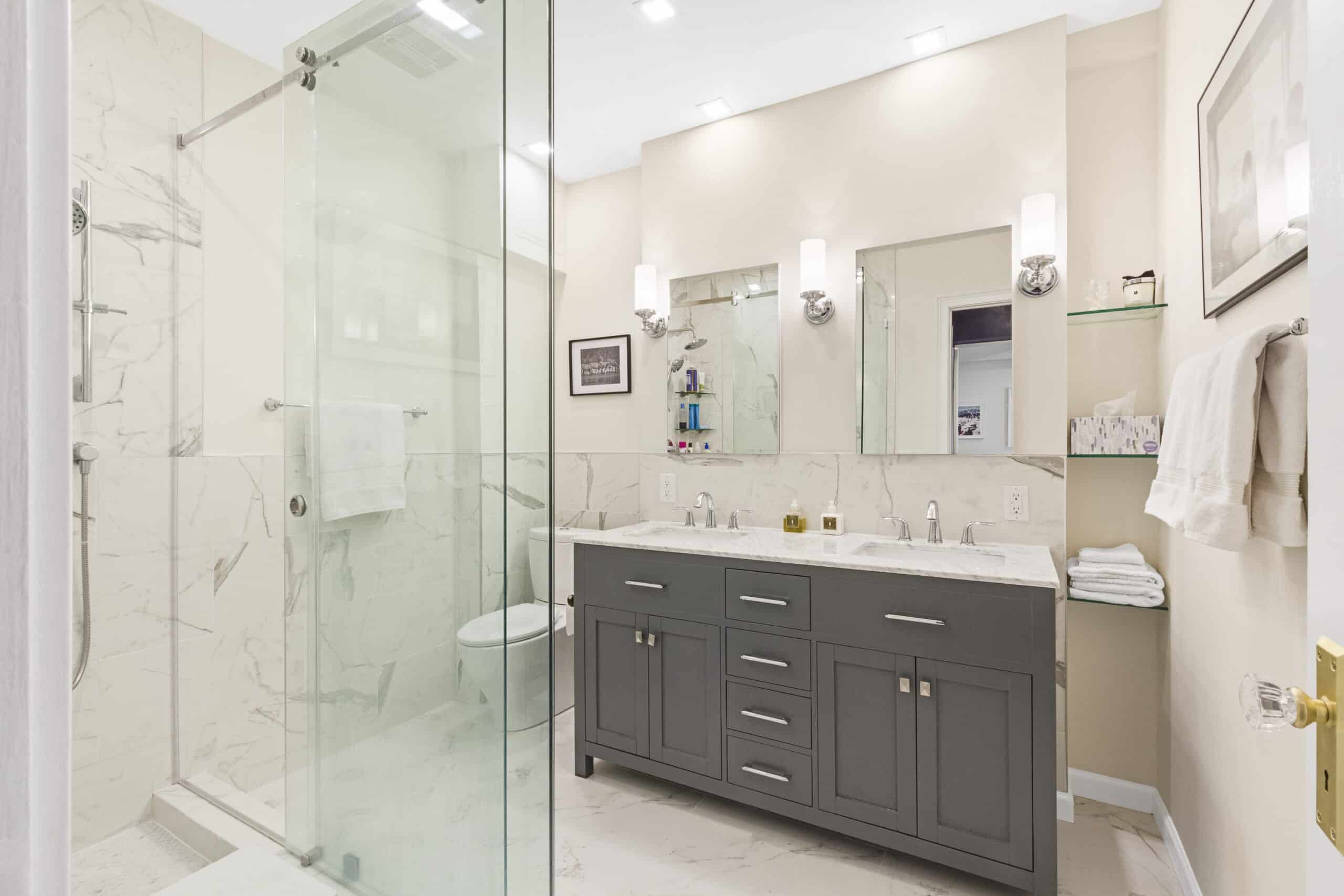 Modern bathroom with a gray double-sink vanity, glass-enclosed shower, and marble tile walls and floor.