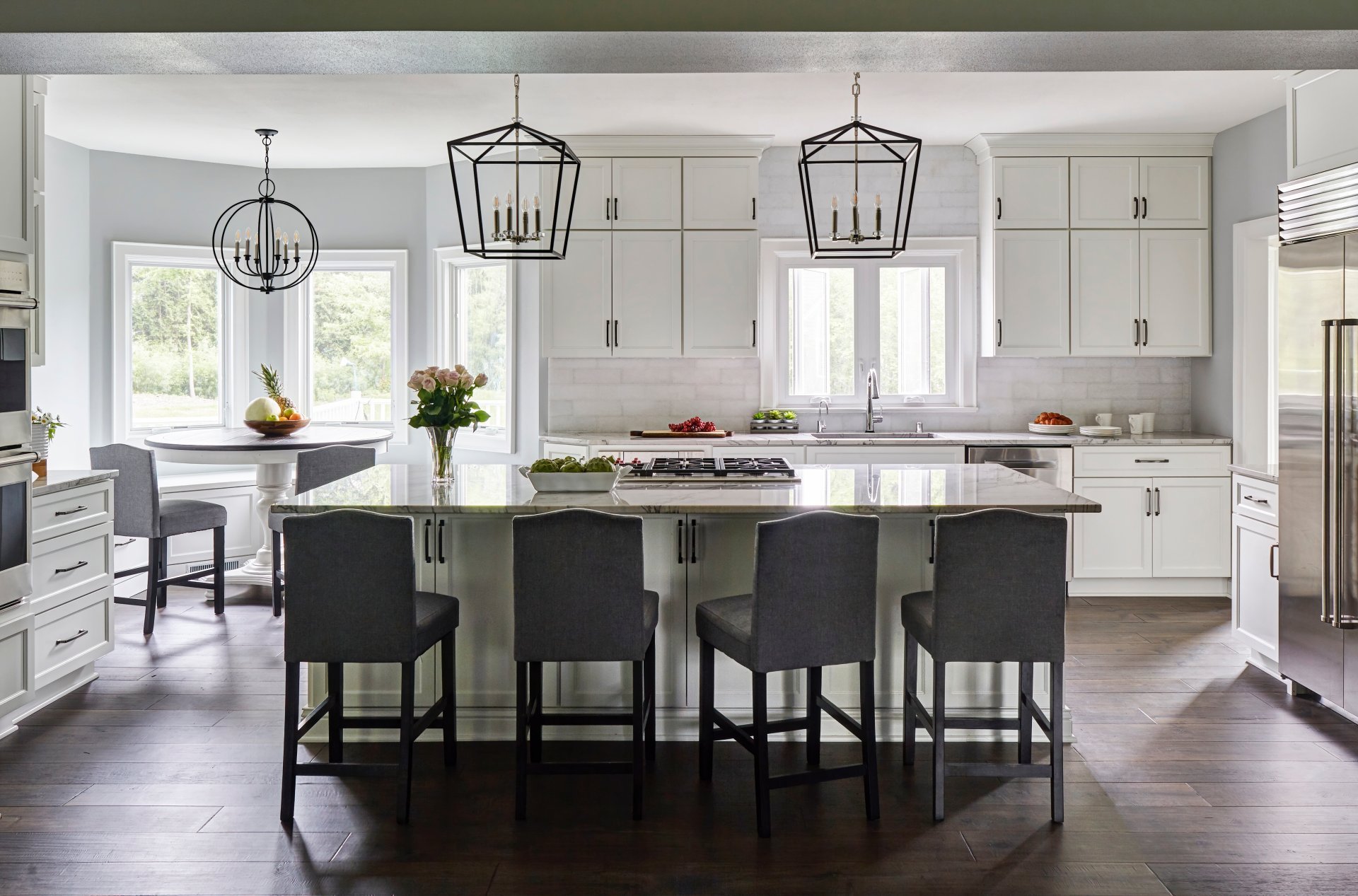 Spacious white kitchen with a large marble island, dark gray bar stools, stainless steel appliances, and geometric pendant lights.