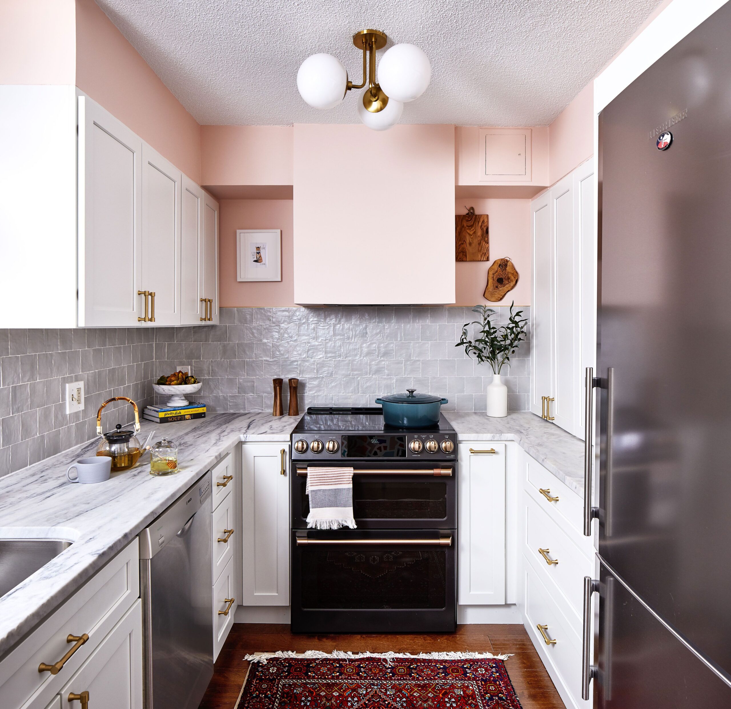 Cozy kitchen featuring soft pink walls, white cabinets with gold handles, marble countertops, and a vintage rug.