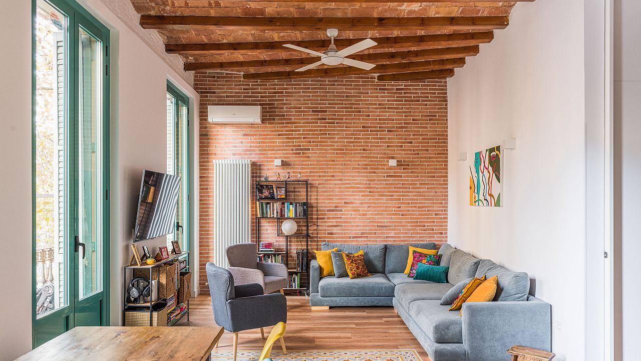 Living room featuring an exposed brick accent wall, wooden beam ceiling, gray sectional sofa with vibrant pillows, and green-framed windows.