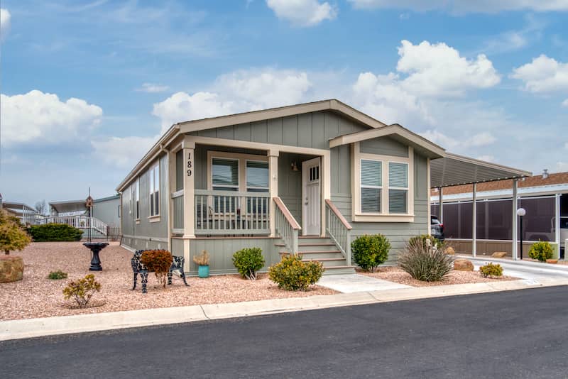 Single-story prefab home with sage green siding, a cozy front porch, desert landscaping, and a carport, situated in a quiet residential neighborhood.