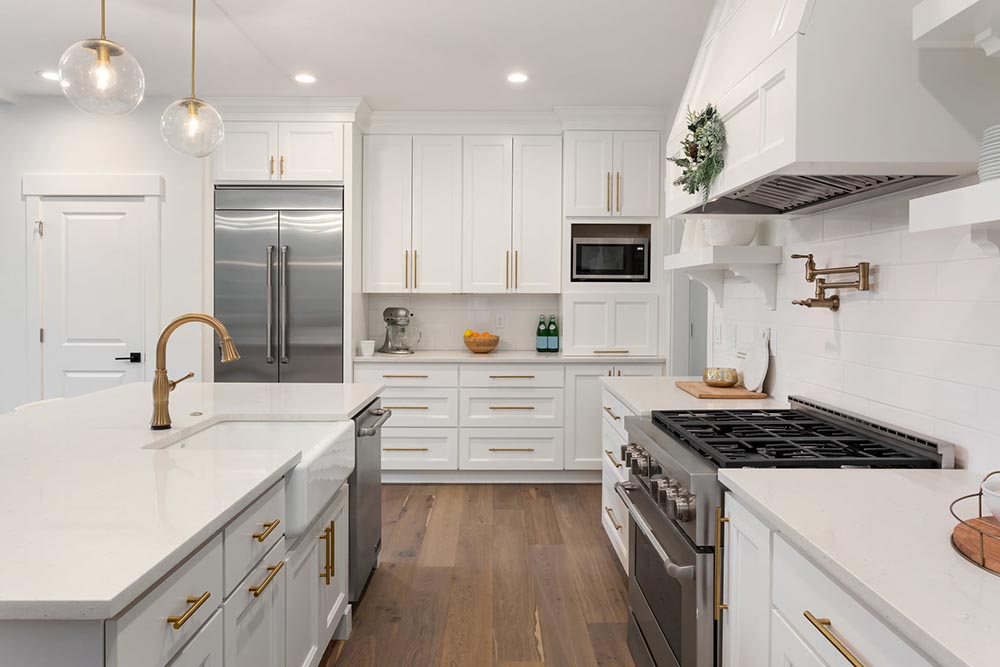 Bright kitchen featuring white shaker cabinets, brass hardware and fixtures, stainless steel appliances, a farmhouse sink, and warm wood flooring.