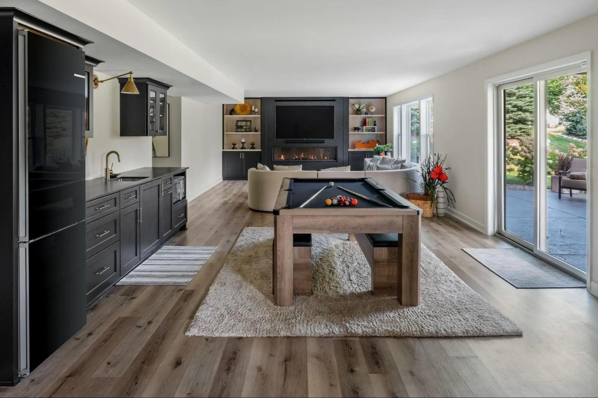 Stylish basement featuring a black kitchenette, pool table, cozy seating area with fireplace, and large glass doors opening to a patio.