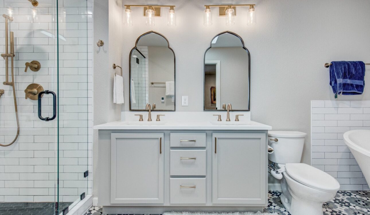 Stylish bathroom with glass shower, double vanity, gold fixtures, and patterned floor tiles.