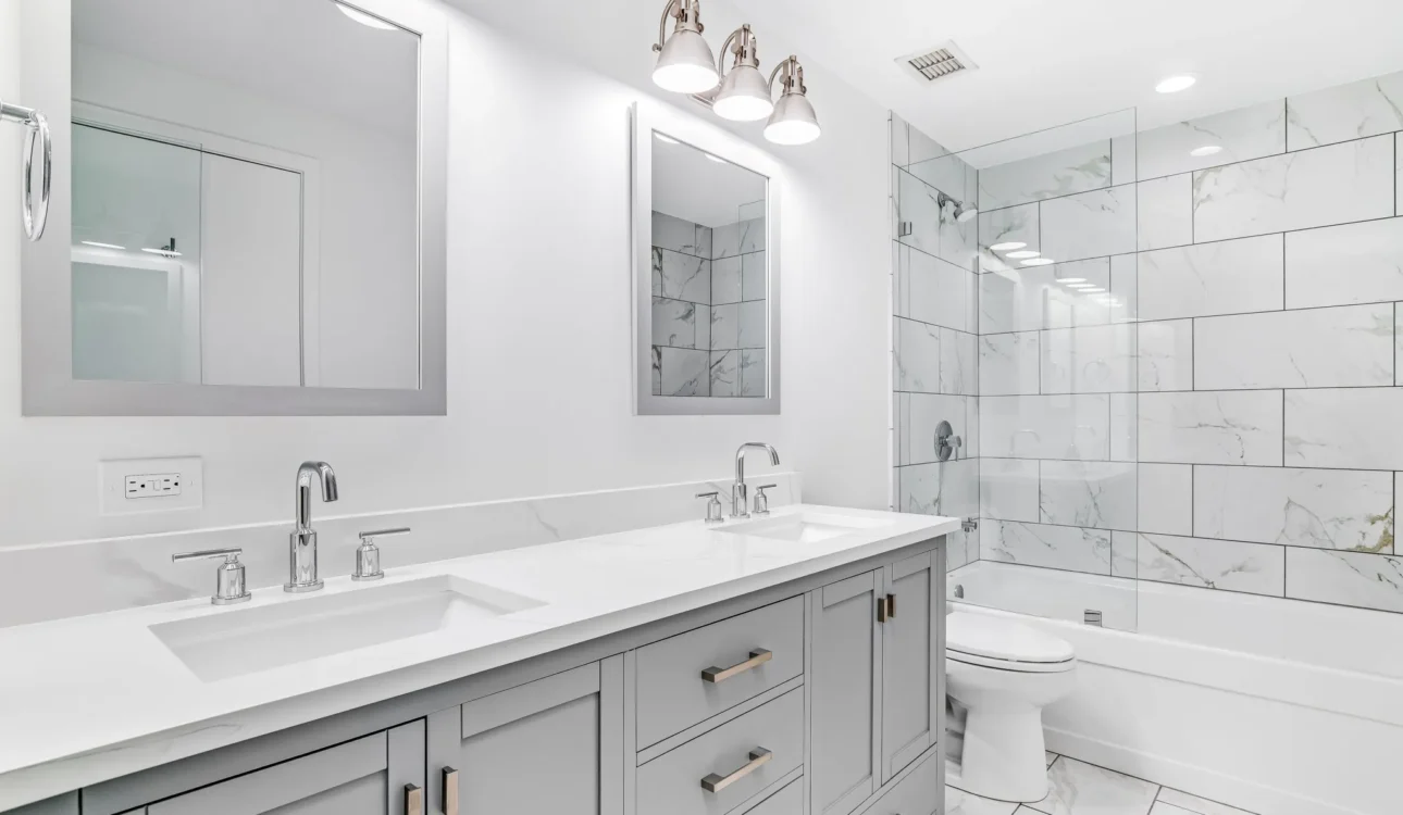 Bright modern bathroom with double vanity, white countertops, glass shower, and marble tile walls.