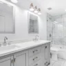 Bright modern bathroom with double vanity, white countertops, glass shower, and marble tile walls.