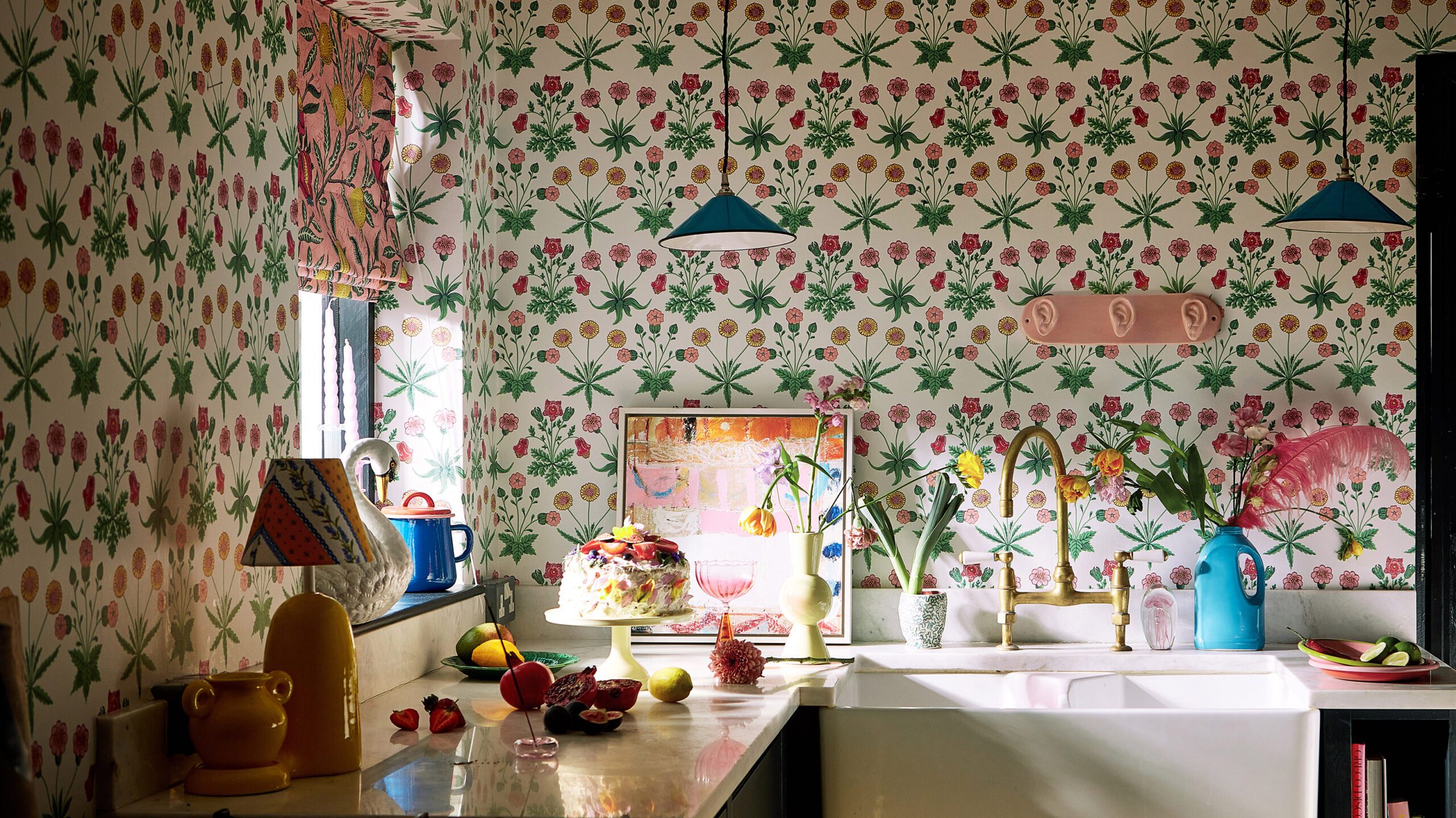 Kitchen with bold floral wallpaper, farmhouse sink, gold faucet, blue pendant lights, and eclectic decor including vases, artwork, and fresh fruit.