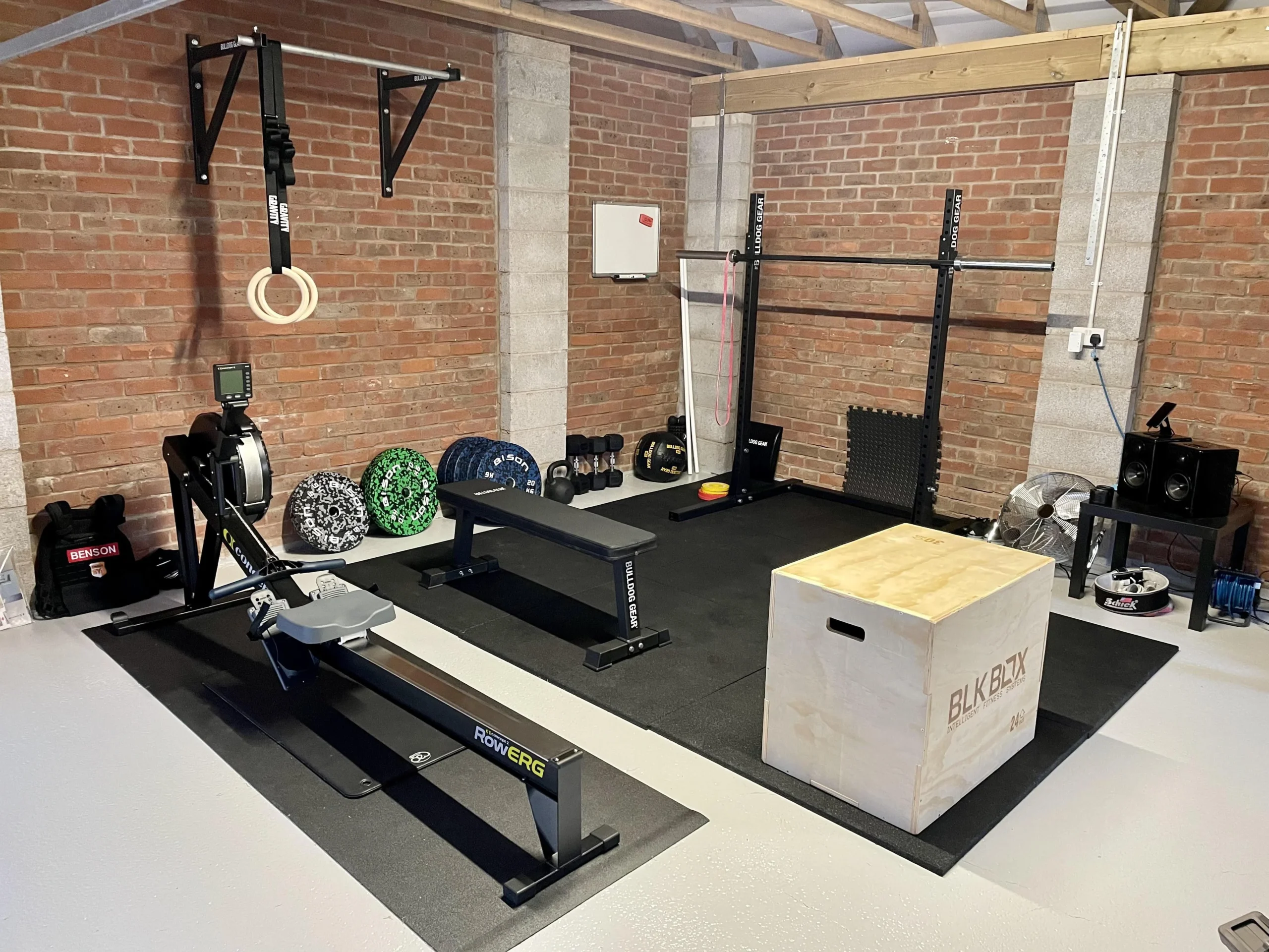 Home gym setup featuring a rowing machine, wooden plyo box, squat rack, weight bench, barbells, bumper plates, and gymnastic rings against a brick wall.