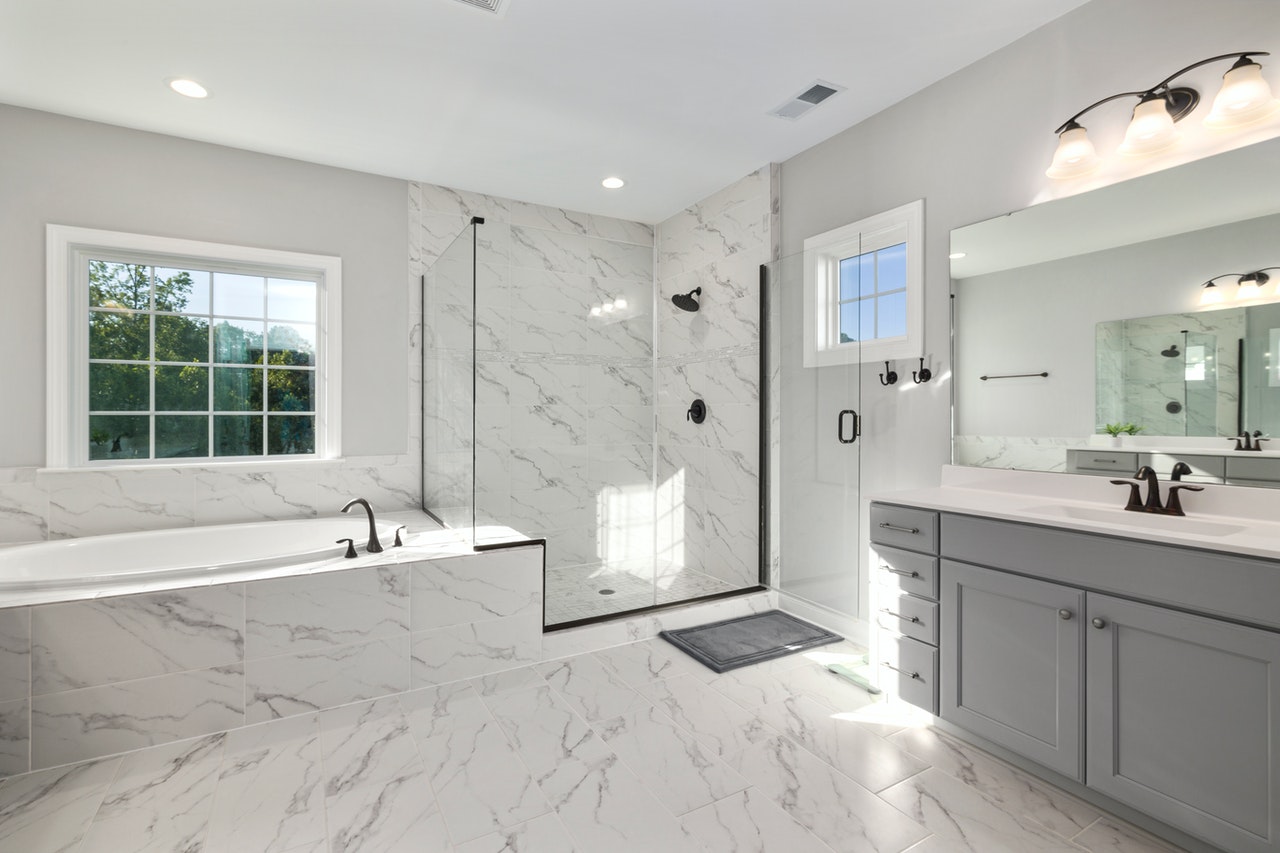 Elegant bathroom featuring marble-look tile floors and walls, a walk-in glass shower, soaking tub by a large window, and a gray vanity with dual sinks.