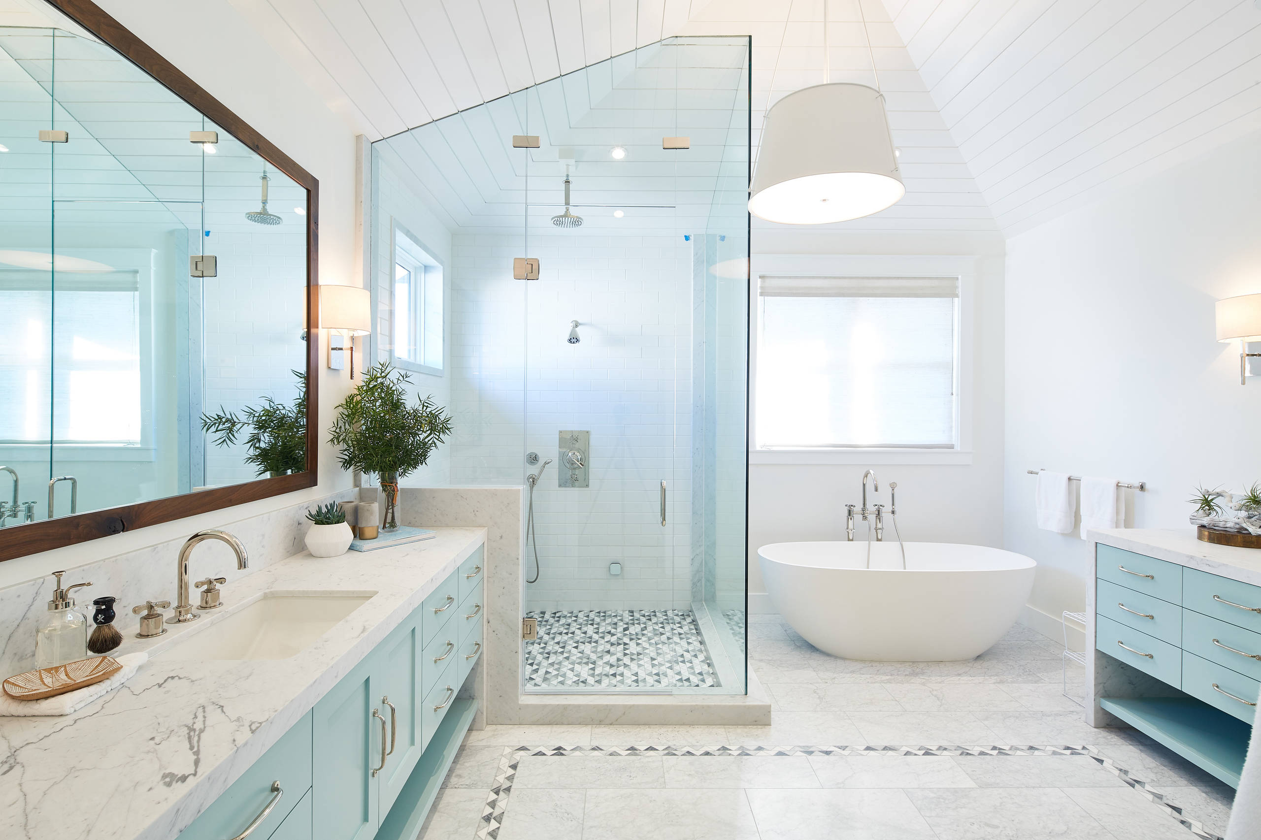 Bright bathroom with glass shower, freestanding tub, aqua vanities, and marble countertops.